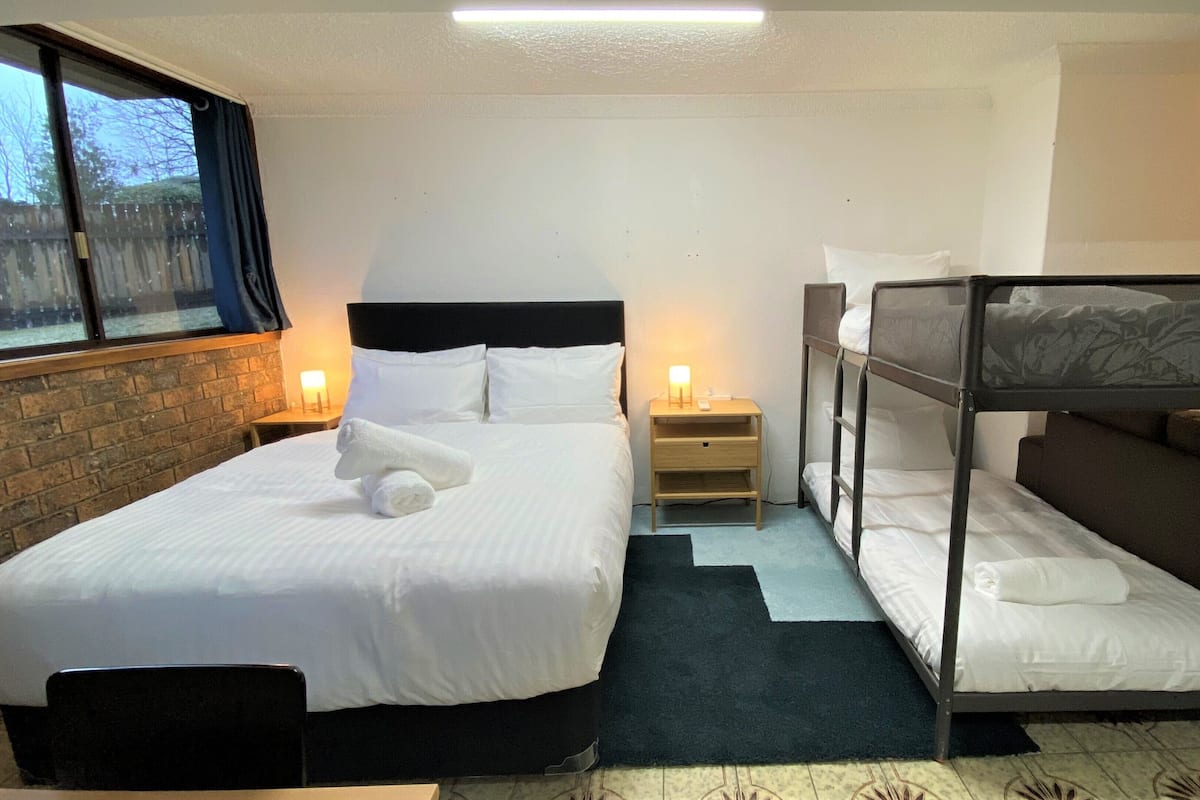 Habitación de hotel con una cama individual, una mesita de noche con lámpara, una cama litera y una ventana con cortinas.
