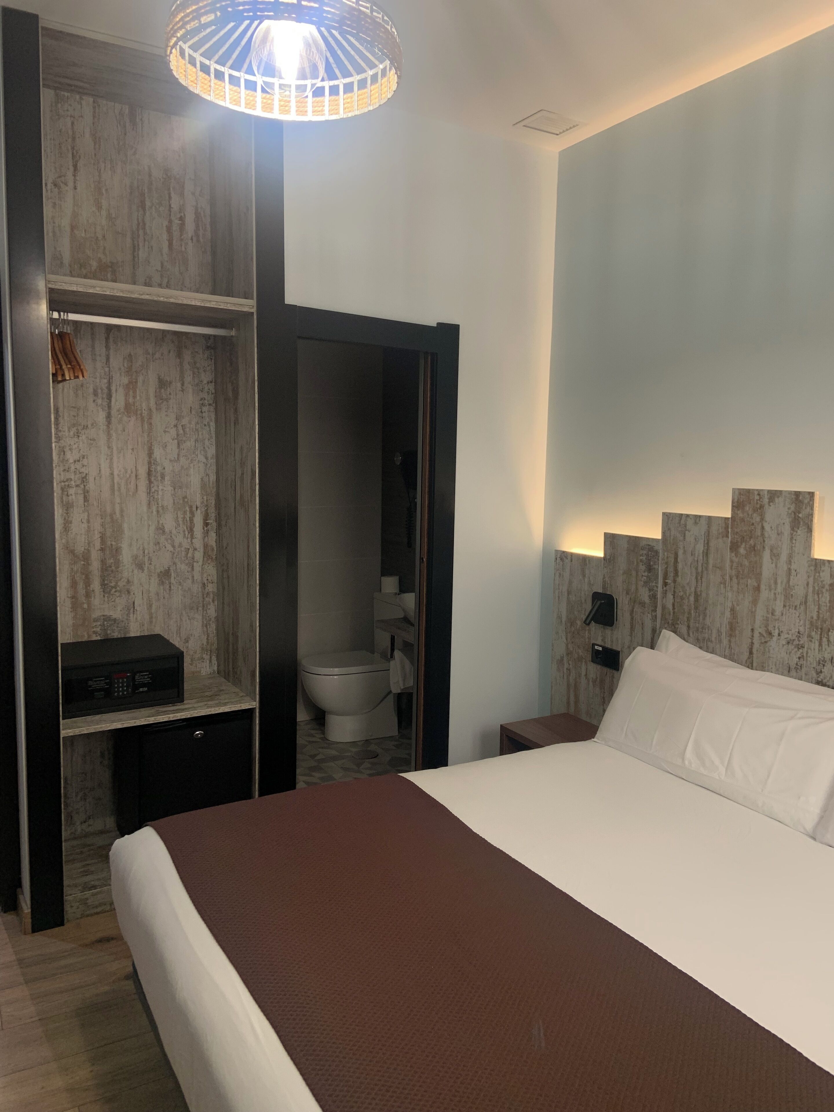 Habitación de hotel con una cama grande, un armario, una mesita de noche, una lámpara y un baño visible a través de una puerta abierta.