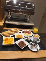 Ein reichhaltiges Frühstücksbuffet mit Grill, Croissants, Rührei, Würstchen, Schinken, Orangenscheiben, Kaffee und einem Glas Orangensaft.