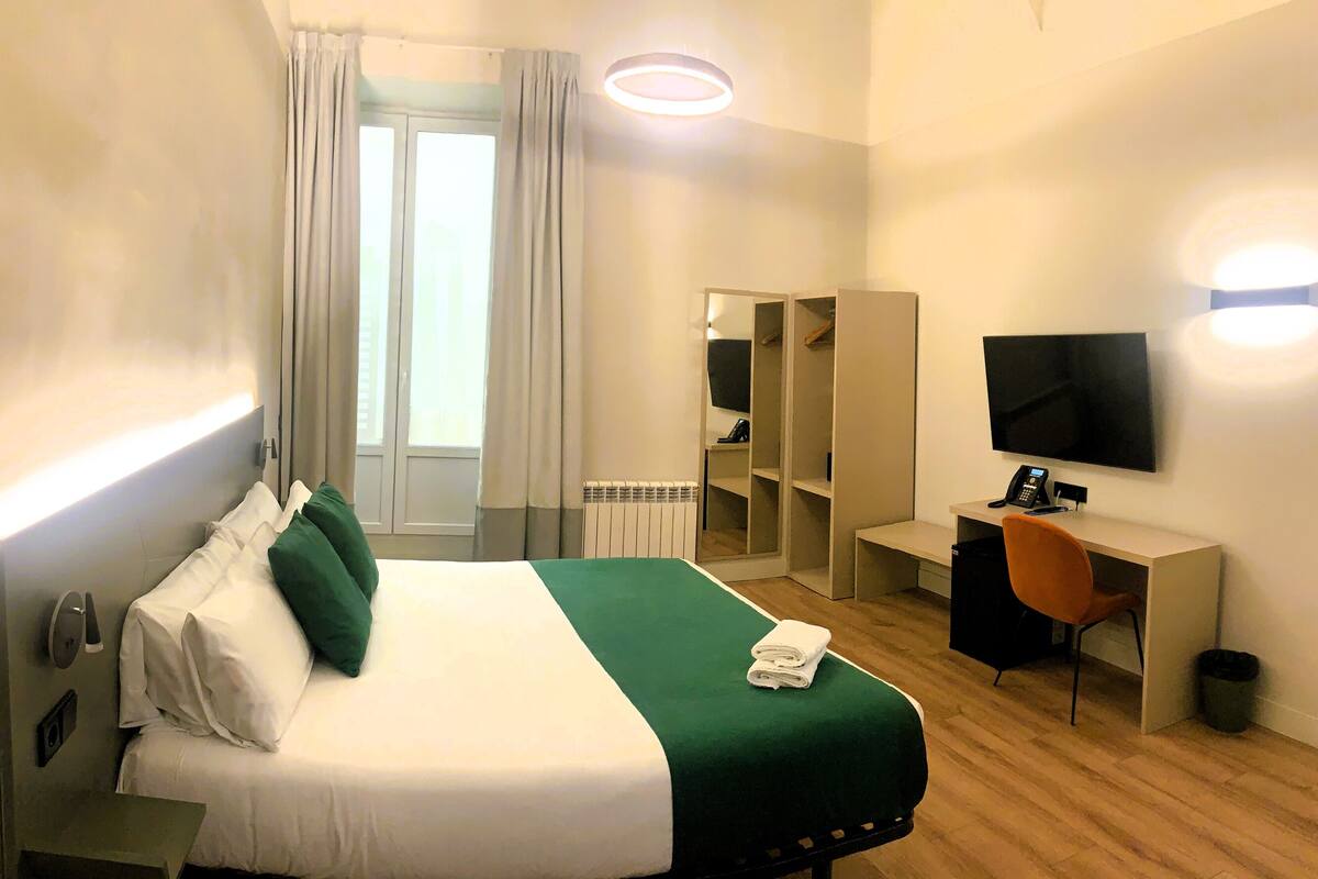 Quarto de hotel com cama, secretária, cadeira, televisão e armário.