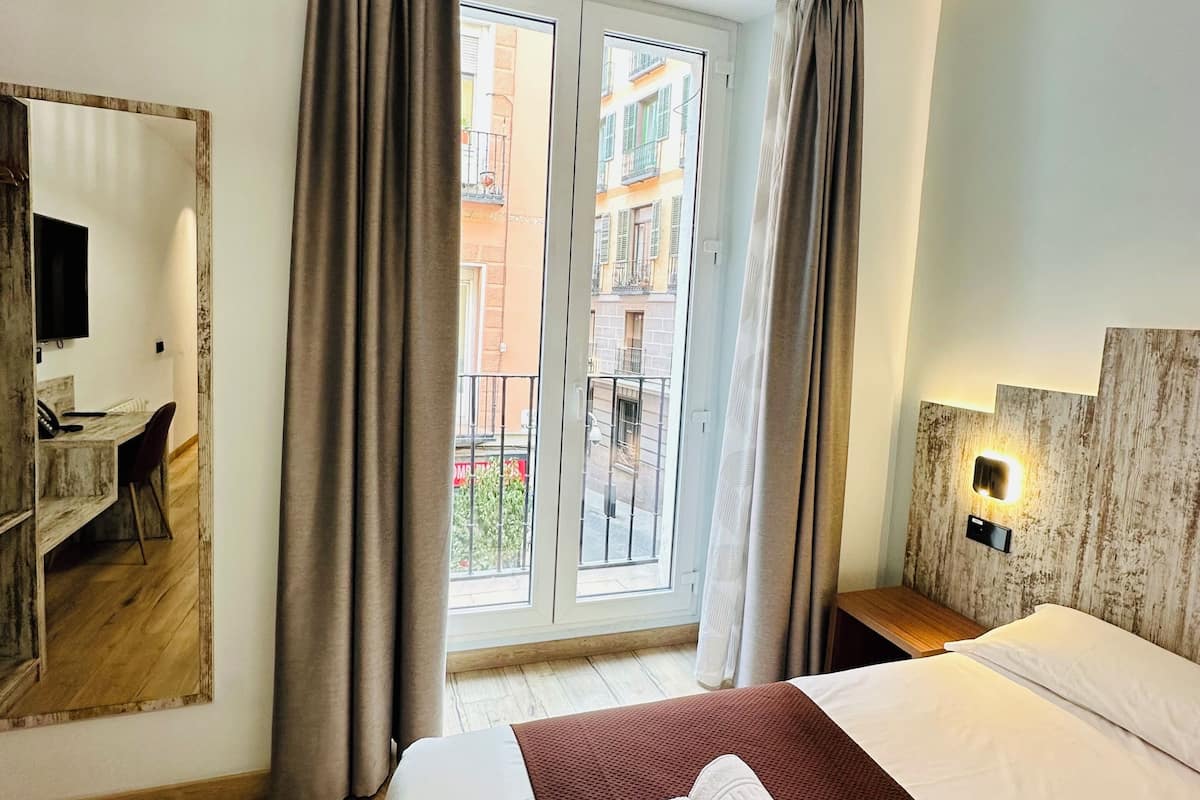 Quarto de hotel com cama, escrivaninha e uma janela grande com cortinas.