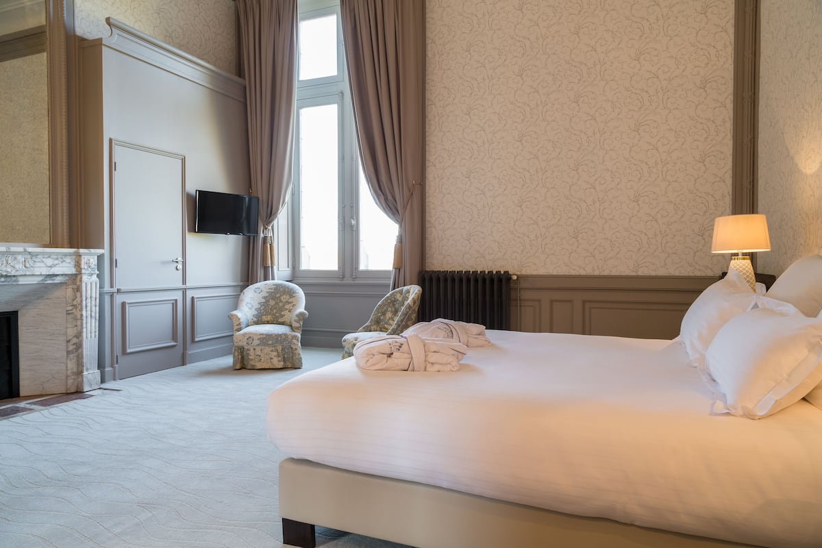A hotel room with a large bed, two armchairs, a fireplace, and a television on the wall.