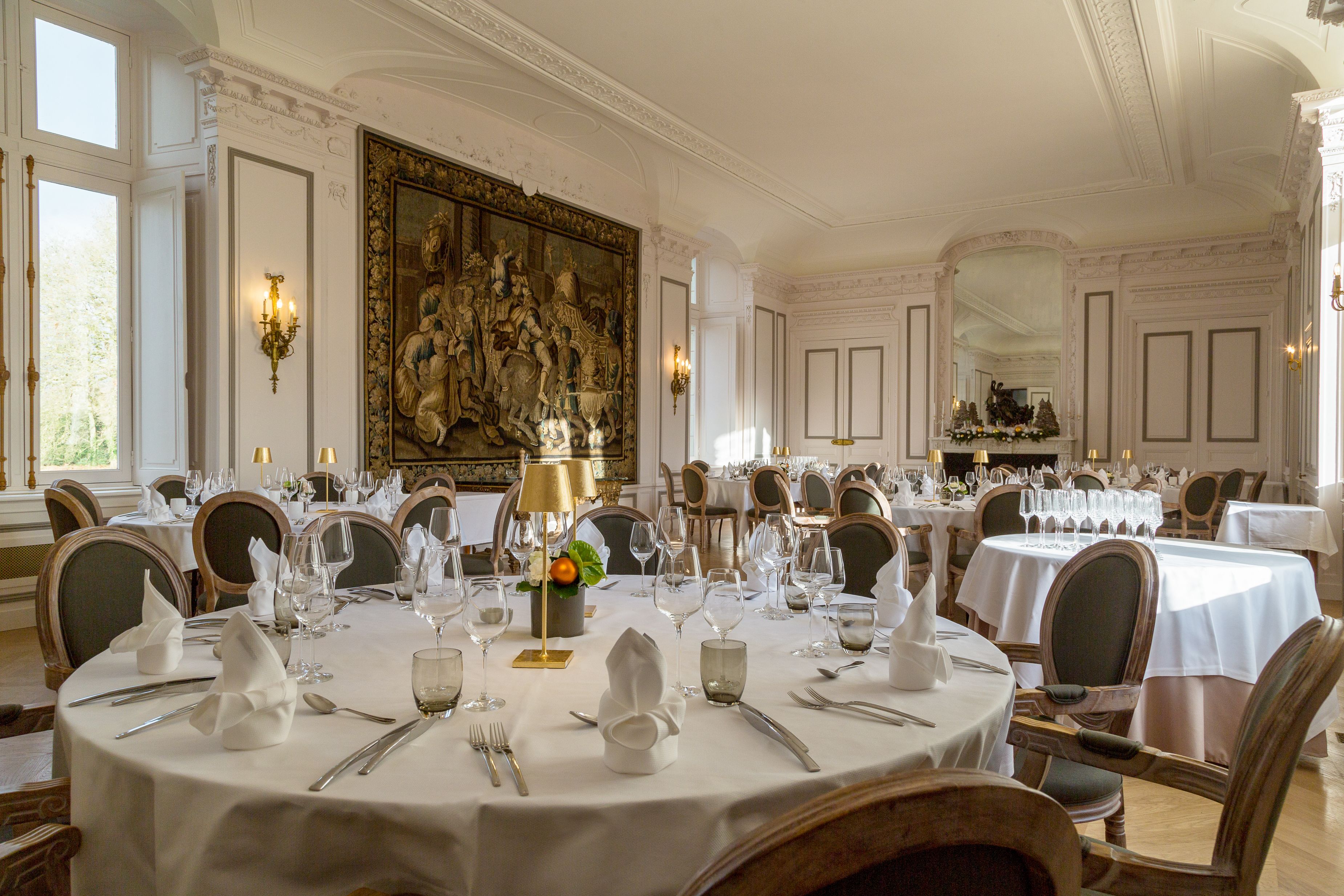 A formal dining room with round tables set for multiple guests, elegant chandeliers, and a large ornate painting on the wall.