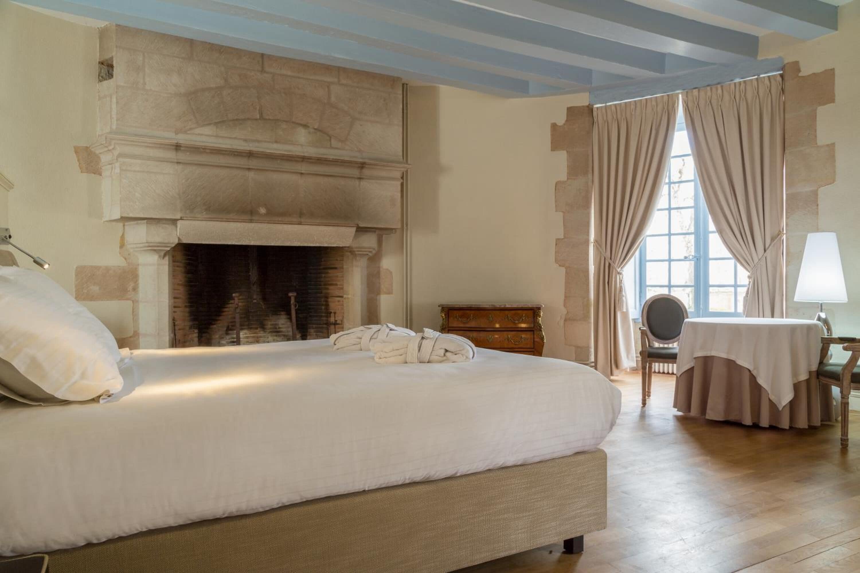 A bedroom with a large bed, a fireplace, a wooden dresser, a table with a chair, and a window with curtains.