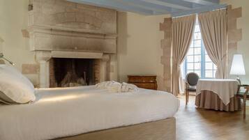 Un dormitorio con una cama grande, chimenea, cómoda de madera, una mesa con una silla y una ventana con cortinas.