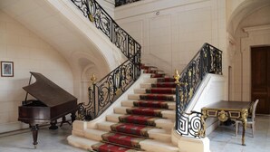 A grand staircase with ornate black railings and red carpeted steps, leading to a landing with a piano and a table.