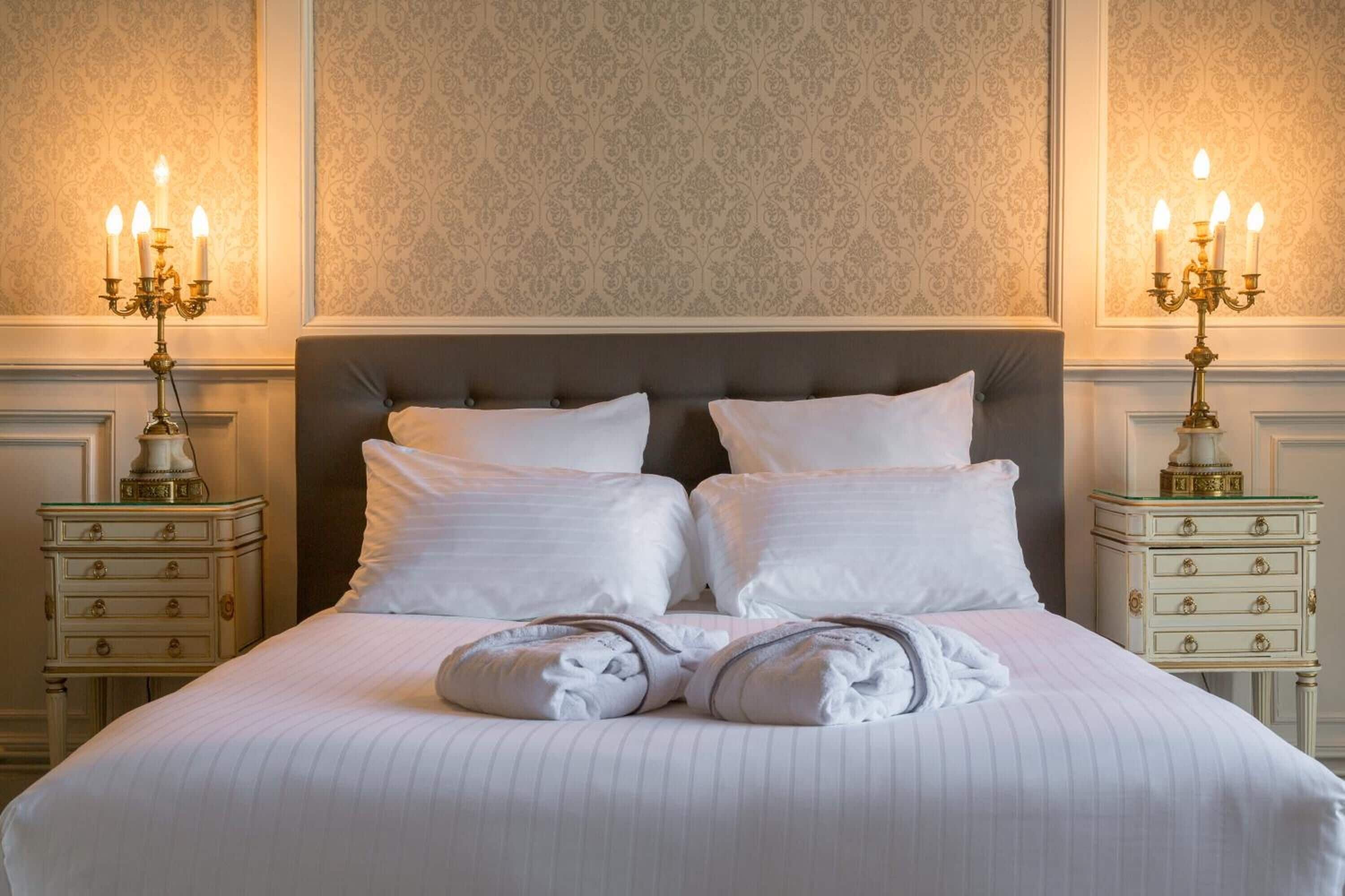 A neatly made bed with white linens and two pillows, flanked by two bedside tables with candelabras.