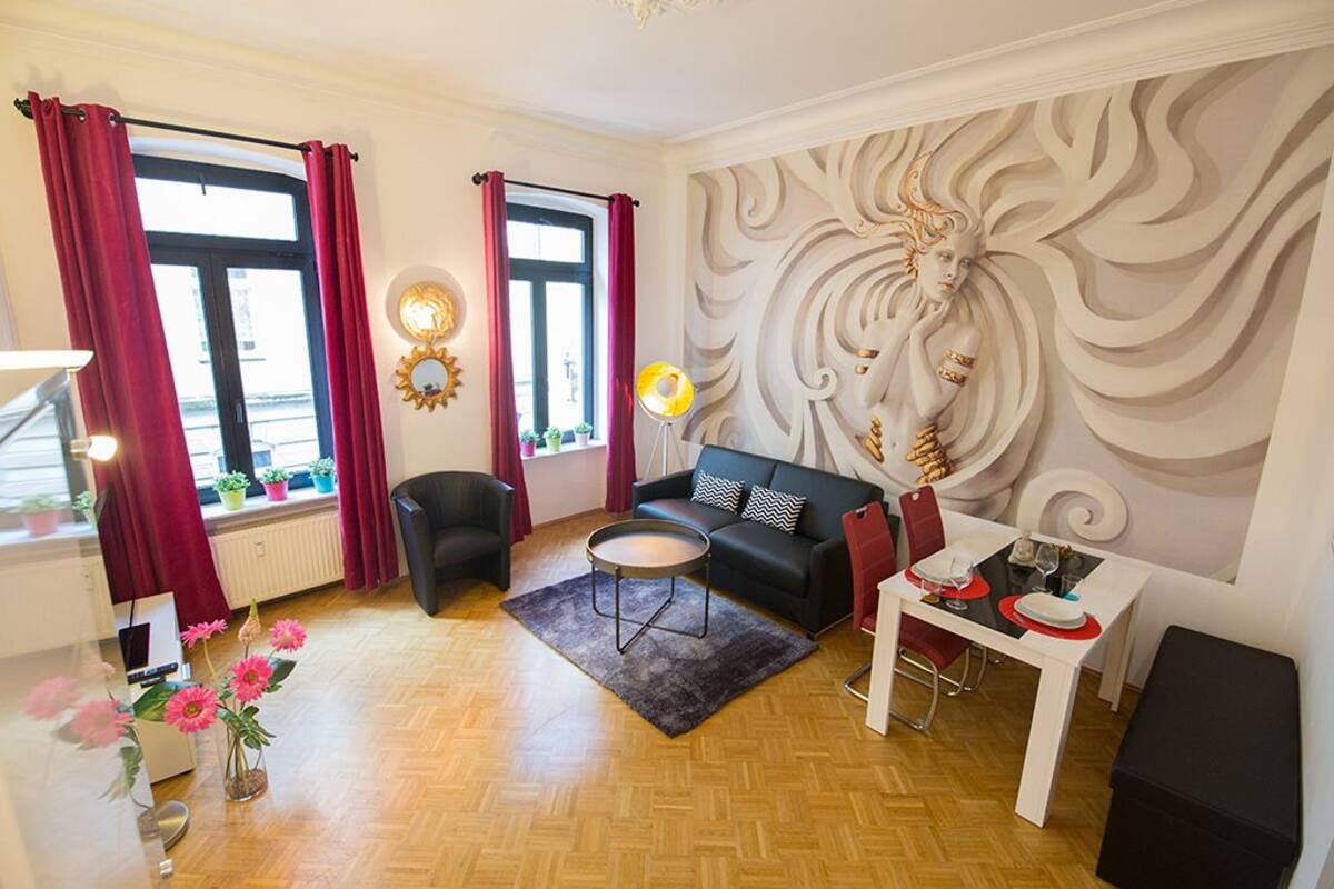 Una sala de estar moderna con un sofá negro, una mesa redonda de centro y una mesa de comedor preparada para dos personas. Hay dos ventanas con cortinas rojas, un mural en la pared y un radiador.