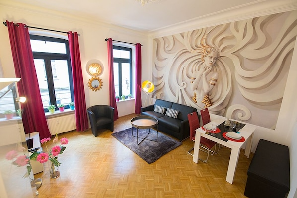 A modern living room with a black sofa, a round coffee table, and a dining table set for two. There are two windows with red curtains, a wall mural, and a radiator.