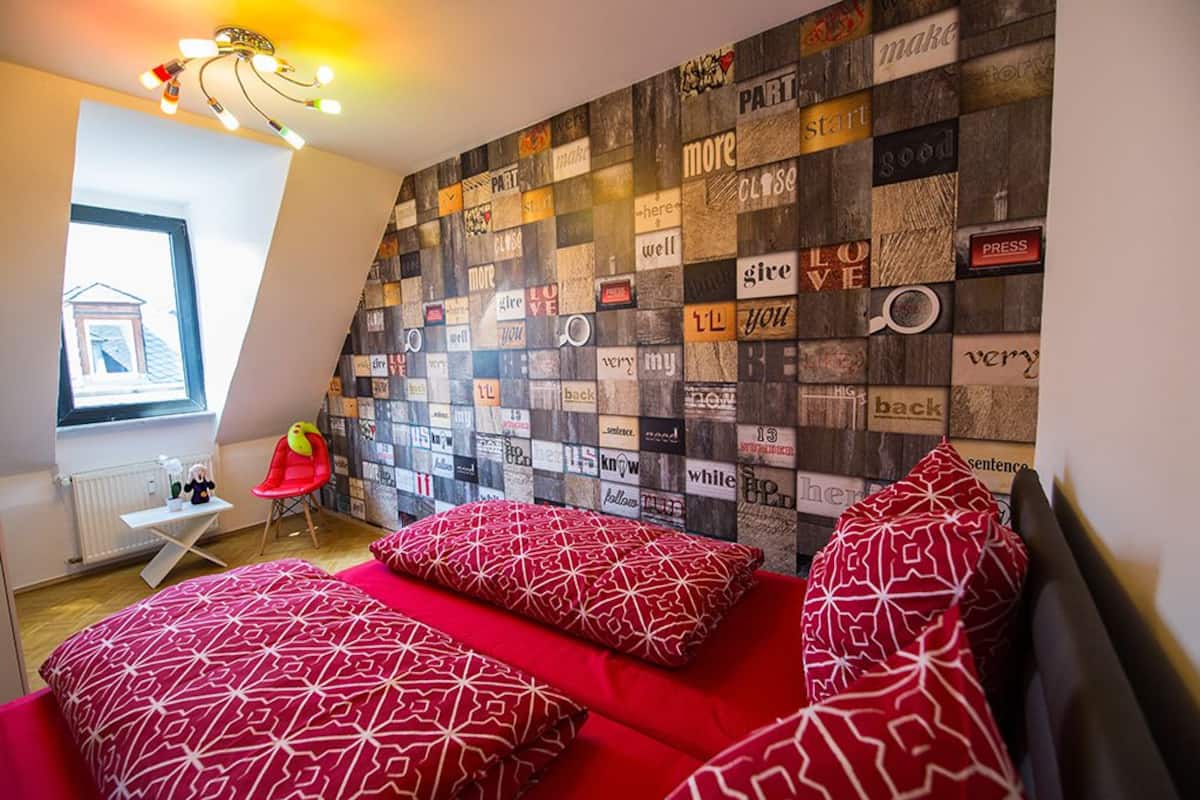 Un dormitorio con techo inclinado, una cama con estampado rojo y una pared decorada con diversas frases.