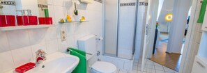 A modern bathroom with a shower, toilet, and sink. There are red and green toiletries on the shelves.