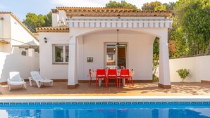 Un área junto a la piscina con un porche cubierto, una mesa de comedor con sillas y sillones de descanso.