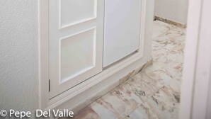 Une armoire blanche dotée d’un réfrigérateur intégré, située dans un couloir aux murs blancs et au sol en marbre.
