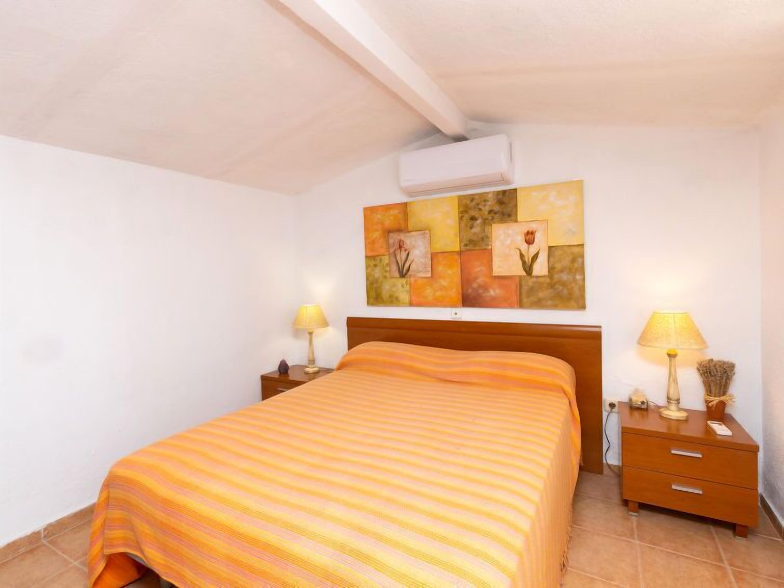 A bedroom with a bed, two bedside tables with lamps, a wall-mounted air conditioner, and a painting above the bed.