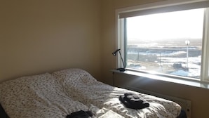 A bedroom with a bed covered in floral bedding, a bedside lamp, and a view of a snowy landscape through a window. - Gljúfraborg (Fjardabyggd)