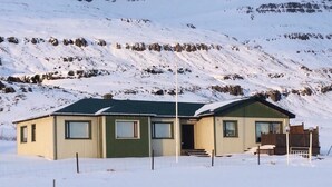 A single-story building with a green roof and a small porch, surrounded by snow-covered mountains. - Gljúfraborg (Fjardabyggd)
