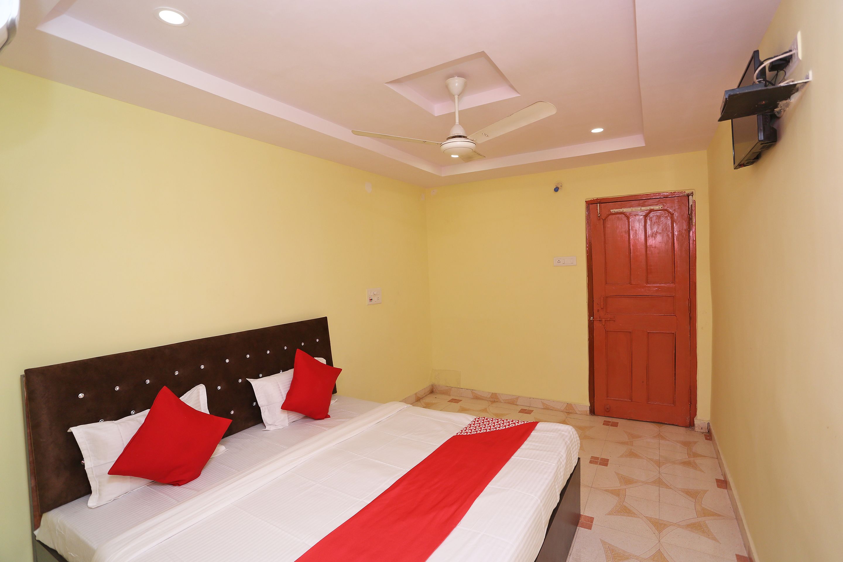 A hotel room with a bed, a ceiling fan, a red door, and a light switch on the wall.
