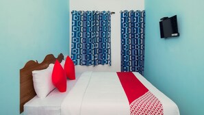 Een slaapkamer met een bed, rode kussens, een witte dekbedovertrek, een rode loper, een blauw patroon gordijn, een airconditioner en een zwarte televisie aan de muur.