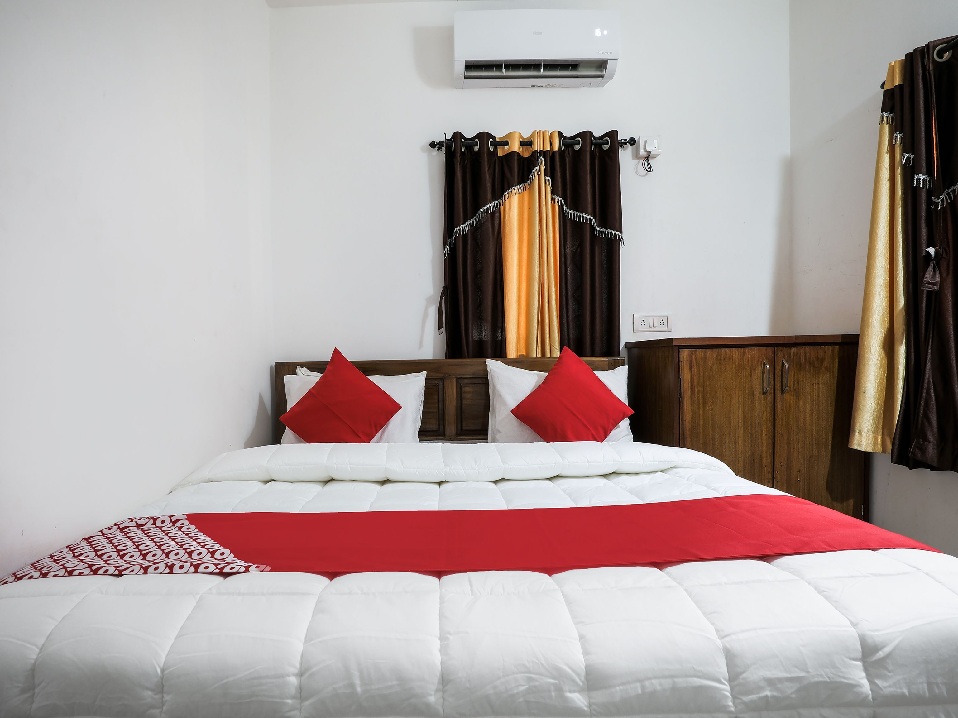 A neatly made bed with white and red bedding, a wooden headboard, and a small wooden cabinet to the right.