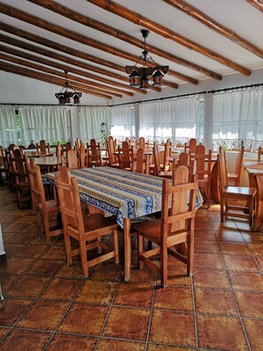 A spacious dining area with wooden tables and chairs, a patterned tablecloth, and a ceiling fan. - BonDia (Rasnov)