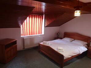 A bedroom with a sloped ceiling, a wooden bed, a nightstand, a lamp, a radiator, and a window with curtains. - BonDia (Rasnov)