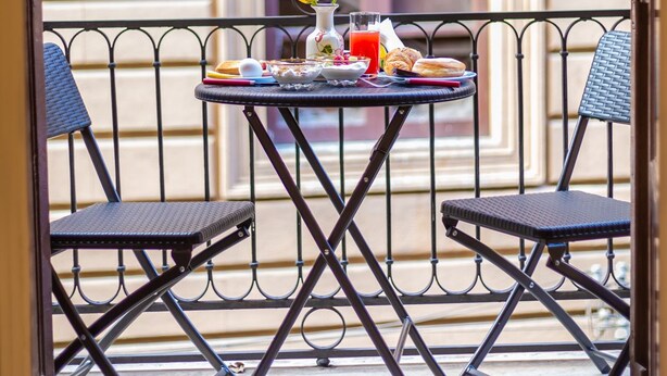 Un balcón con una mesa lista para el desayuno, dos sillas y una ventana con persianas.