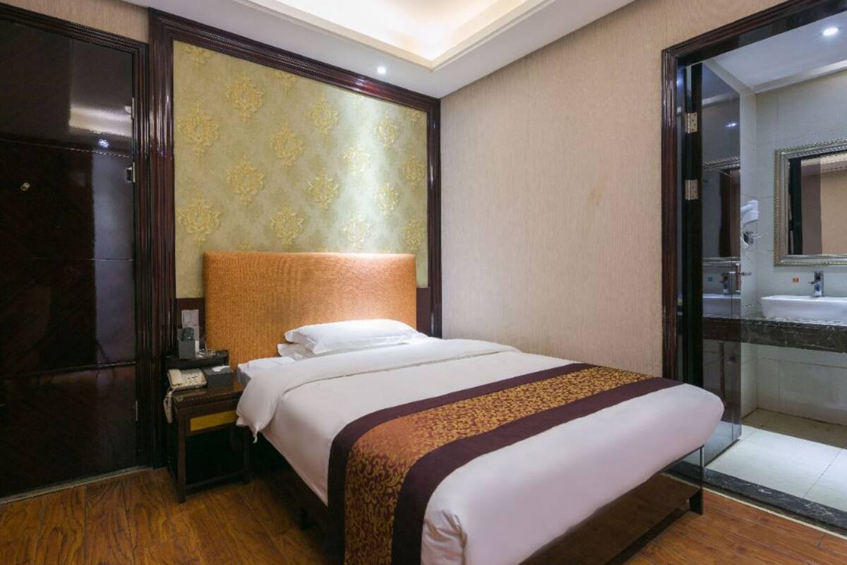 A hotel room with a large bed, a nightstand, a wooden floor, a patterned headboard, and a bathroom with a sink and mirror.