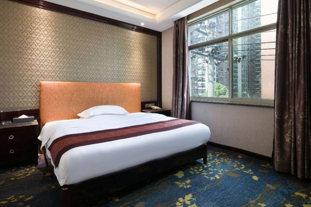 A hotel room with a large bed, a nightstand, a window with curtains, and a patterned carpet.