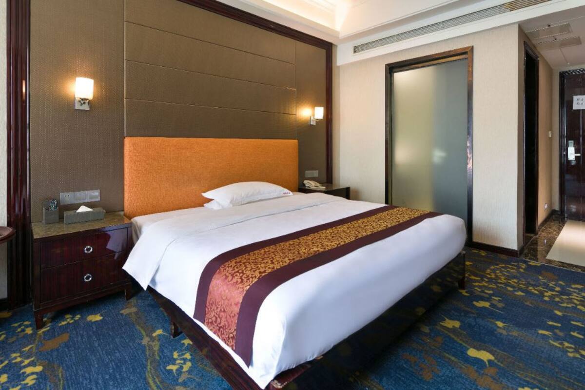 A hotel room with a large bed, a wooden headboard, a nightstand, a bedside table, a mirror, and a carpeted floor.