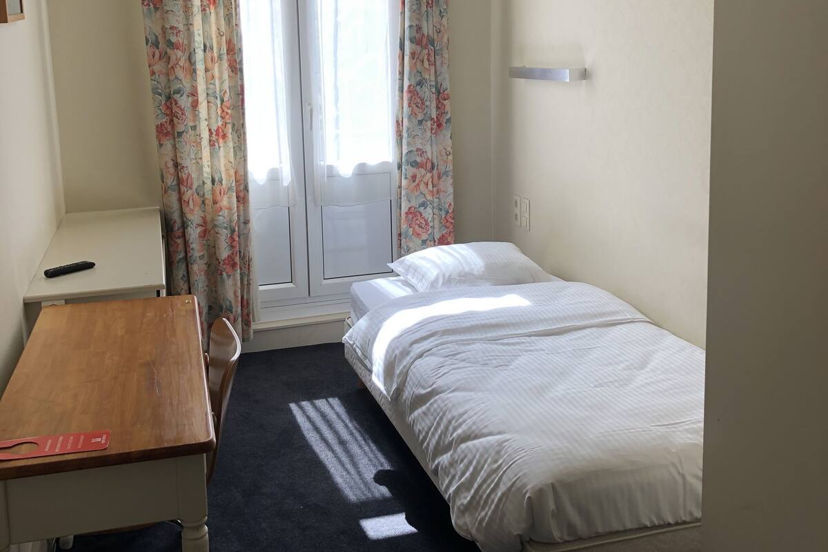 A small, well-lit room with a single bed, a desk, and a chair.