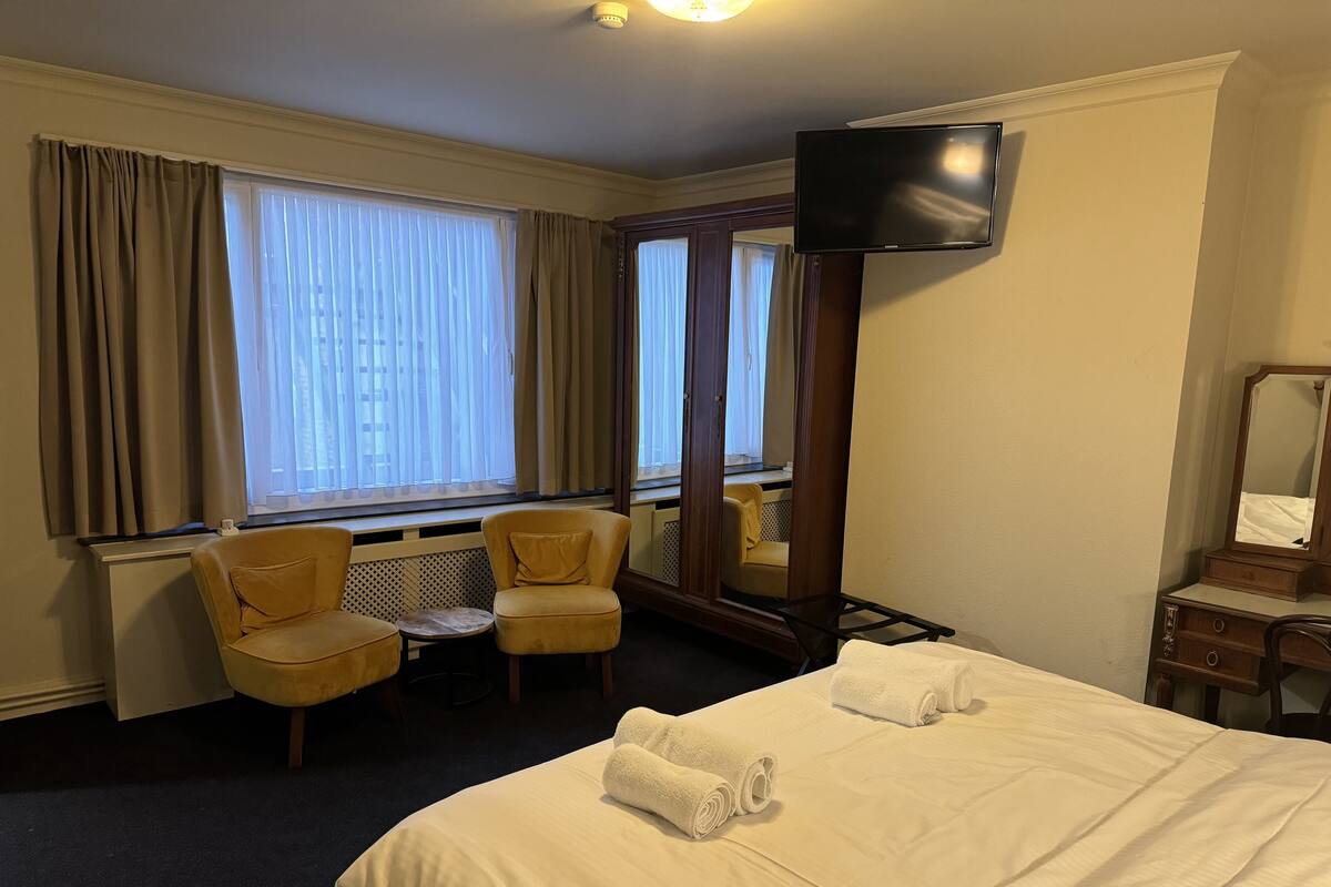 A hotel room with a bed, two armchairs, a small table, a TV, and a mirror.