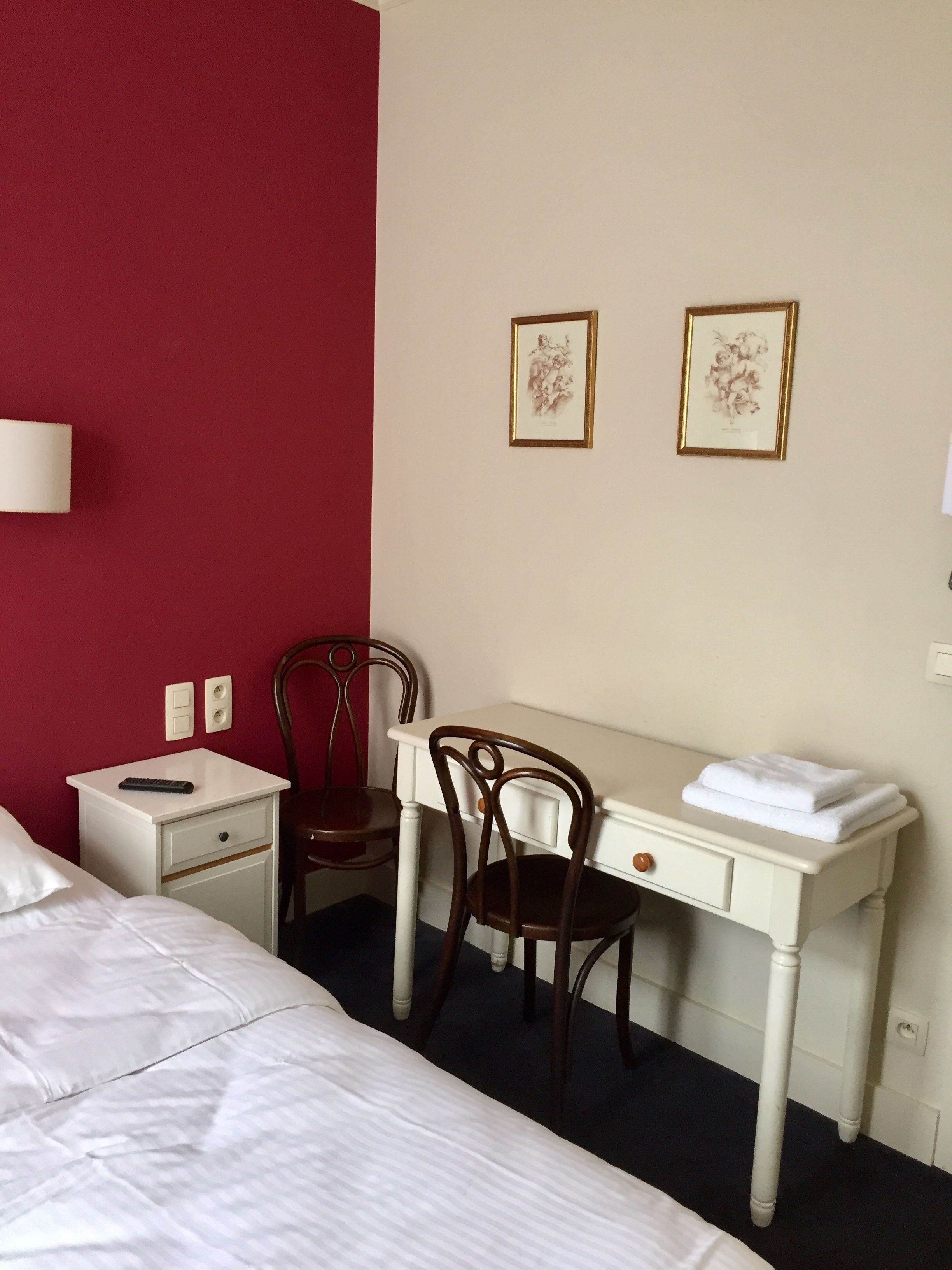 A hotel room with a bed, a desk with a chair, a nightstand, and two framed pictures on the wall.