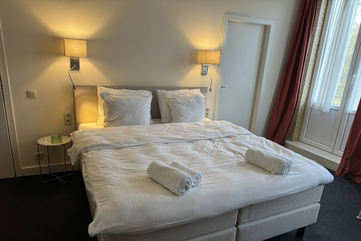 A neatly made bed with white linens and two rolled towels, a bedside table with a lamp, and a window with curtains.