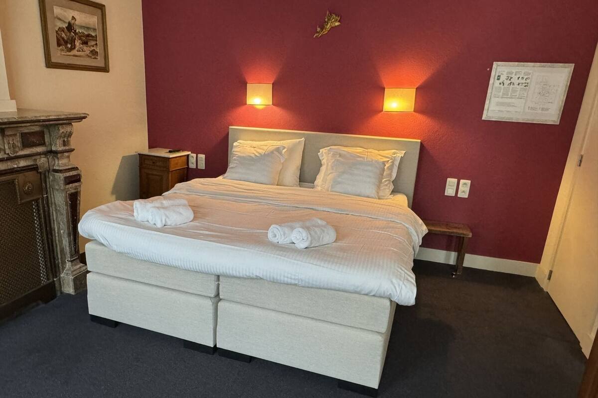 A neatly made bed with white linens and towels, a wooden nightstand, a small table, and a wall-mounted light fixture.