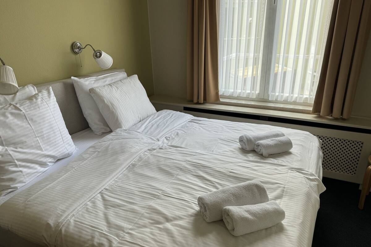 A neatly made double bed with white linens and two rolled towels, a bedside lamp, and a window with curtains.