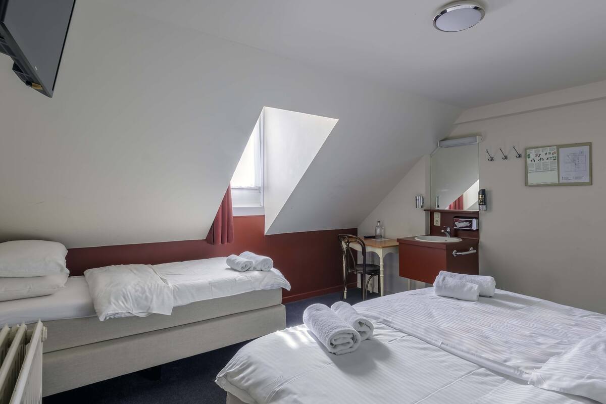 A hotel room with two beds, a desk, a chair, a small bathroom, and a window providing natural light.