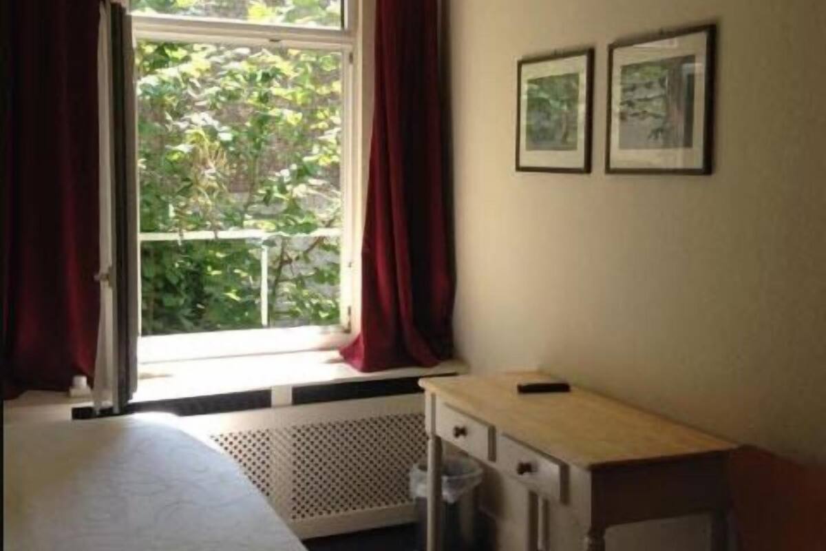 A bedroom with a bed, a desk, and a chair. There is a window with red curtains and two framed pictures on the wall.