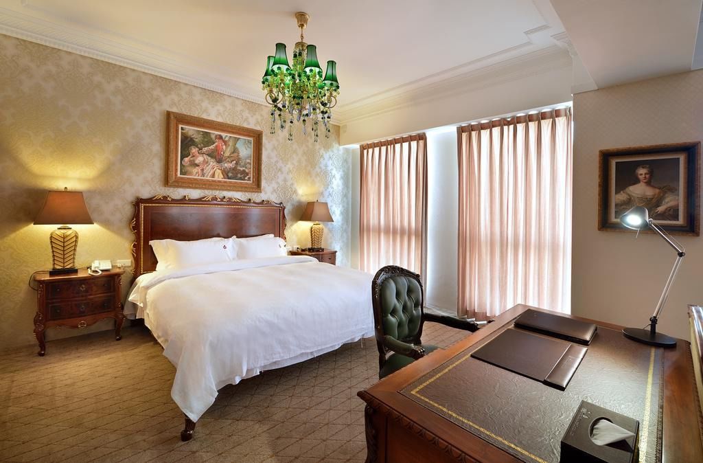 Una habitación de hotel con una cama grande, dos mesitas de noche con lámparas, un escritorio con una silla, una araña y cortinas.