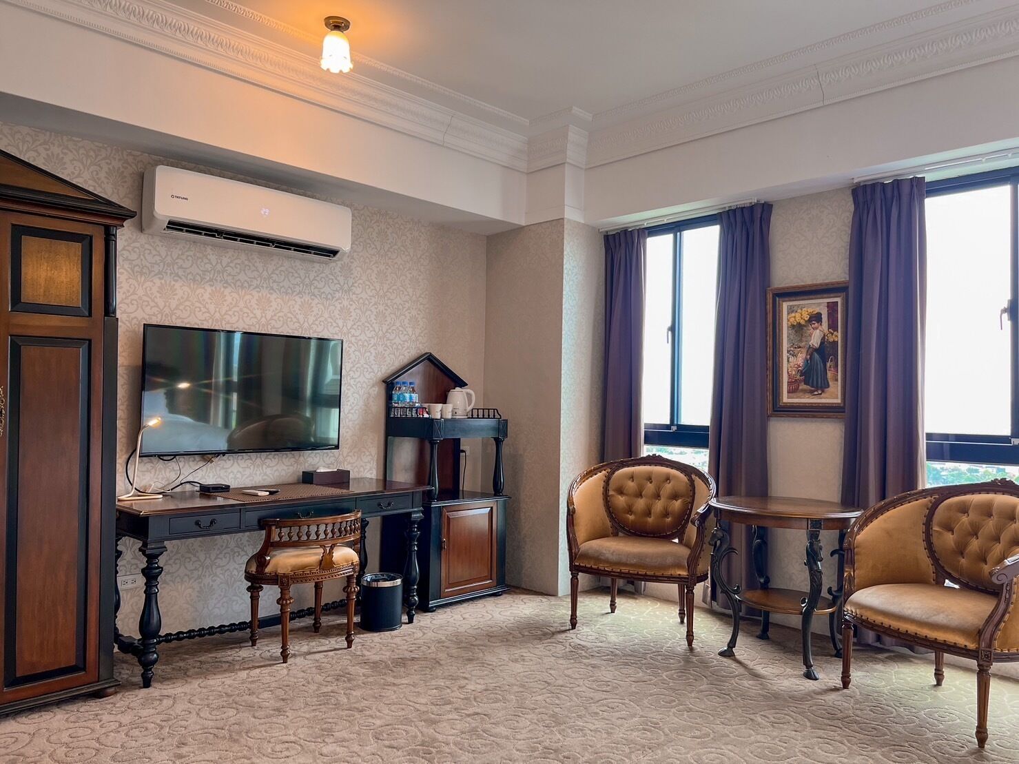 A hotel room with a large flat-screen TV, a desk with a chair, two upholstered chairs, a small table, and a decorative picture on the wall.