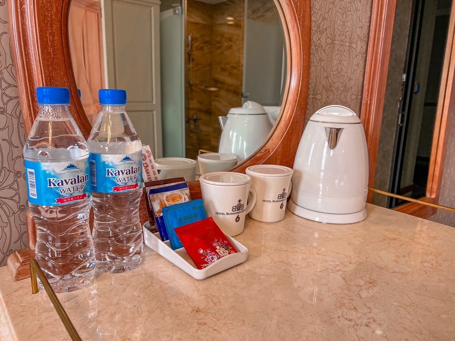 A hotel room counter with two bottles of Kavalon water, a tea set, a white kettle, and a tray with a red envelope.