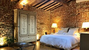 Une pièce avec un mur en briques, un plafond en bois, un lit avec une literie blanche, une table de chevet avec une lampe, une armoire aux portes décoratives et une petite table sur laquelle est posée une plante.