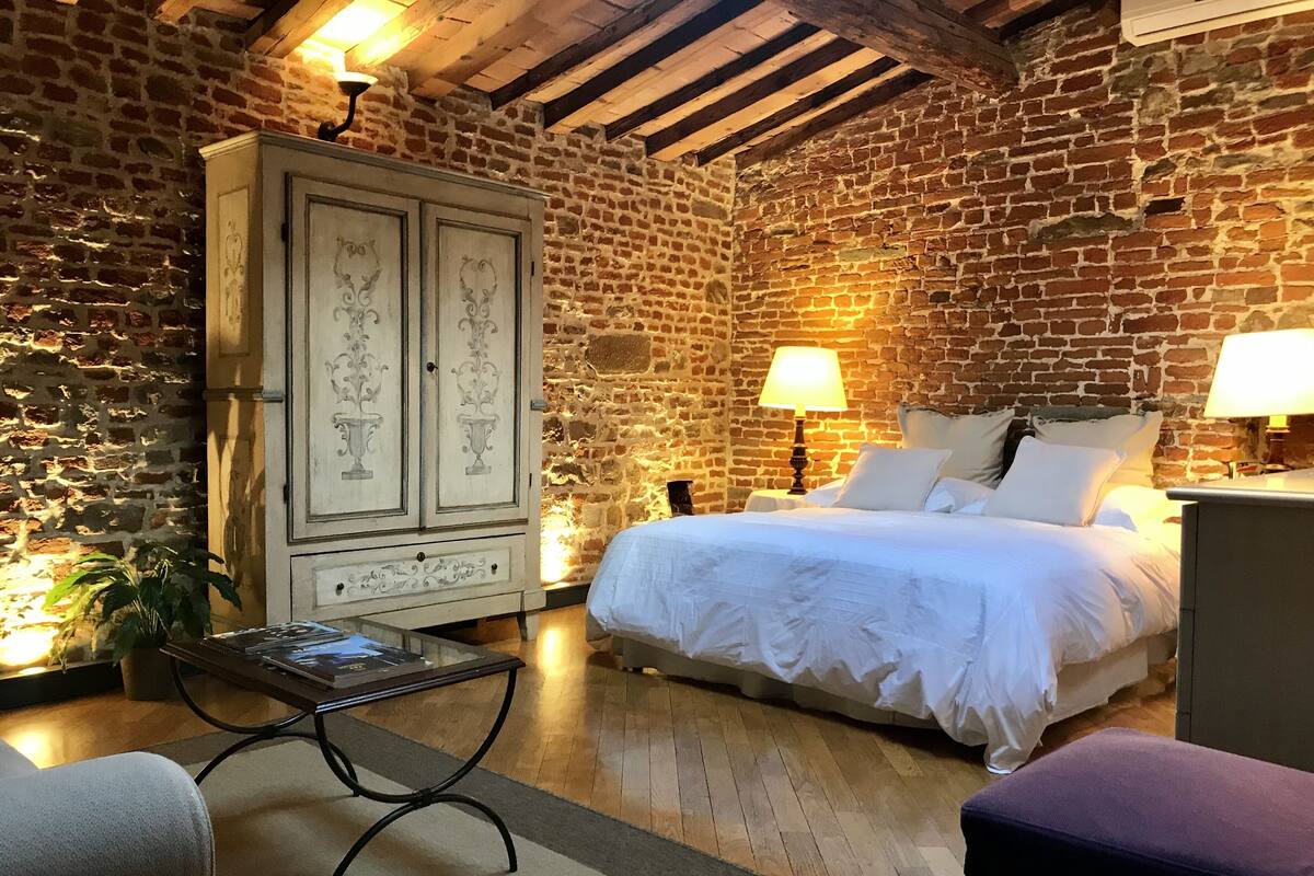 Quarto com parede de tijolos, teto de madeira, cama com roupa de cama branca, mesa de cabeceira com abajur, armário com portas decorativas e uma pequena mesa com uma planta.
