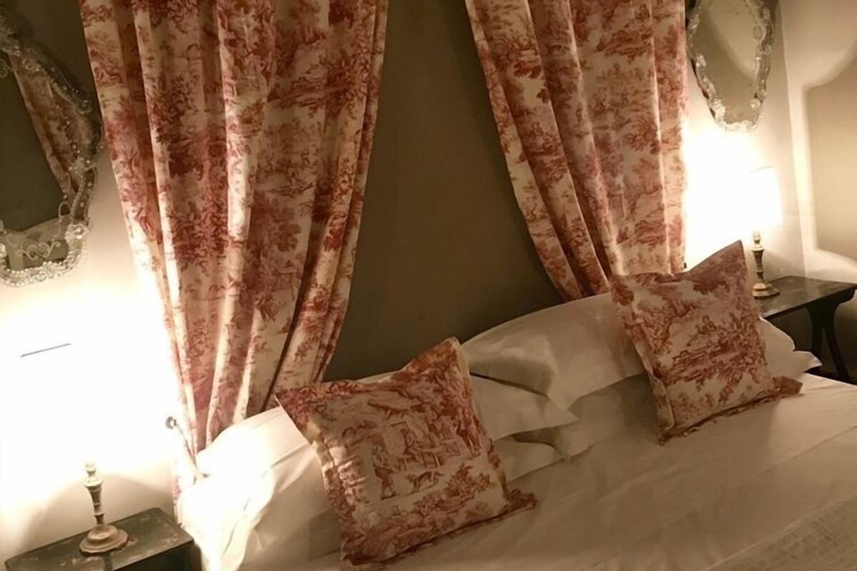 Quarto com cama, cortinas florais, almofadas a combinar, mesa de cabeceira e espelho.