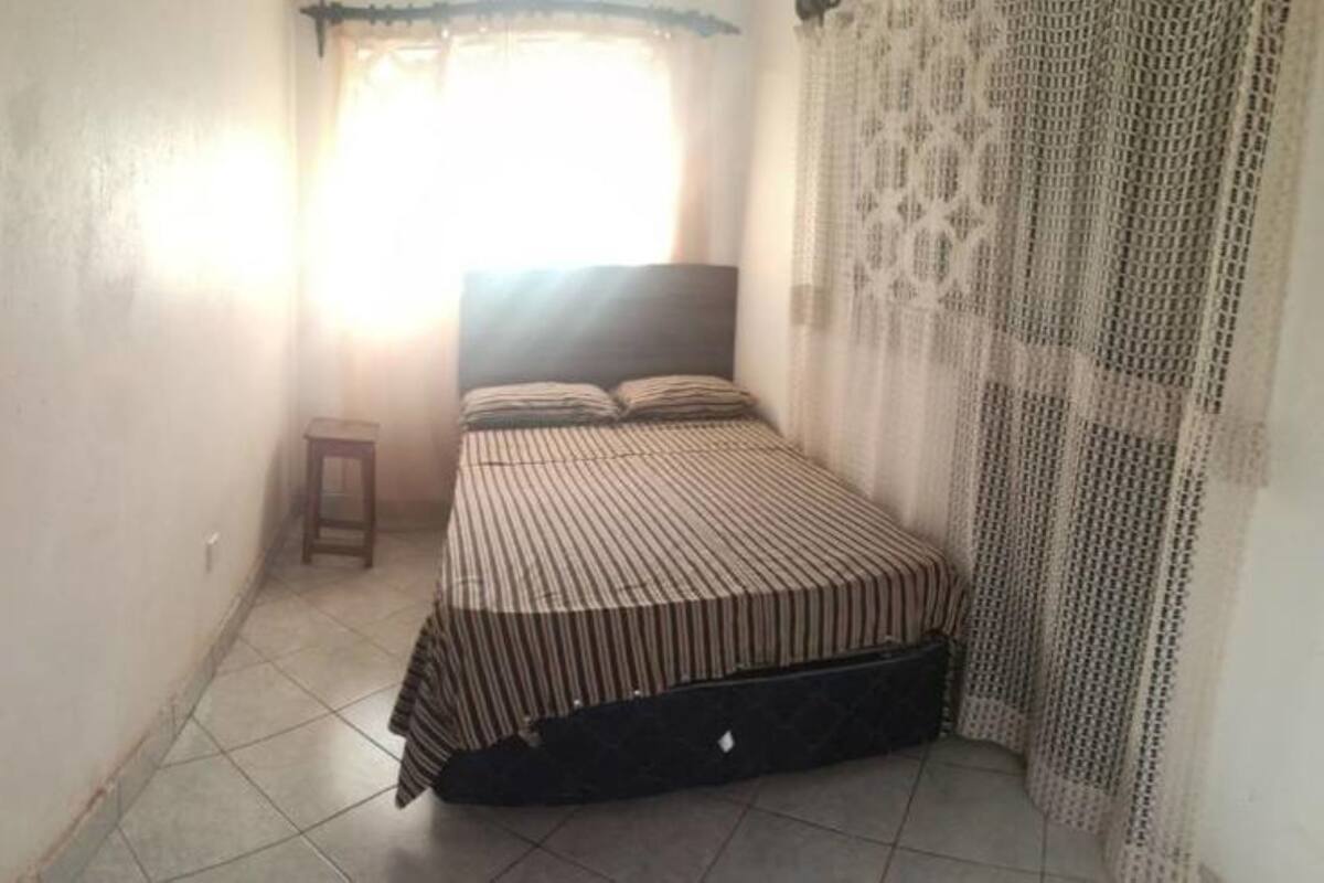 Un dormitorio con una cama, un taburete pequeño y una ventana con cortinas.