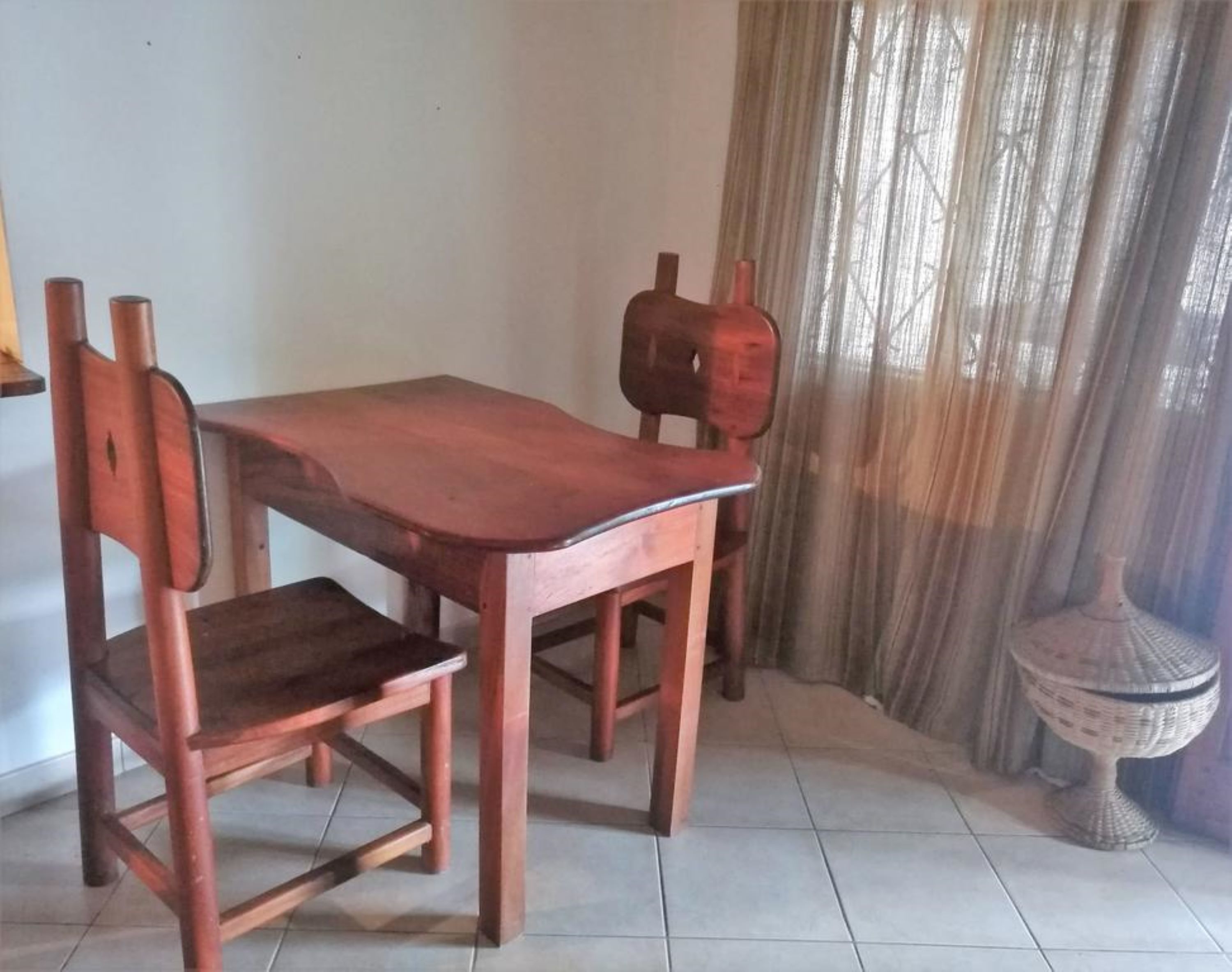 Een houten eettafel met vier stoelen, een betegelde vloer en een raam met dunne gordijnen.