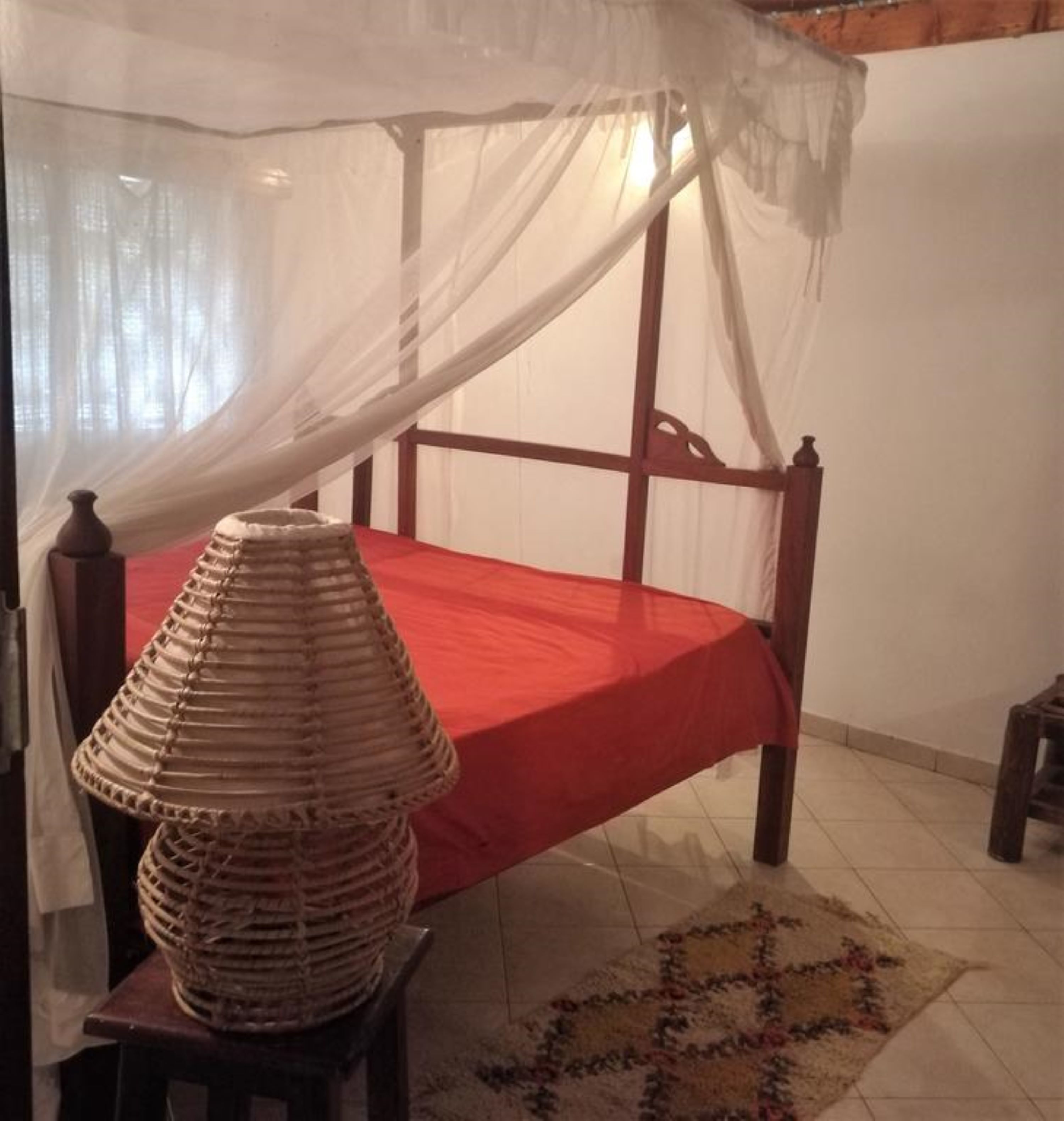 Un lit recouvert d’une courtepointe rouge et surmonté d’un baldaquin blanc, une lampe en osier posée sur un tabouret en bois, et un tapis à motifs sur le sol.