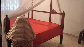 Un lit recouvert d’une courtepointe rouge et surmonté d’un baldaquin blanc, une lampe en osier posée sur un tabouret en bois, et un tapis à motifs sur le sol.