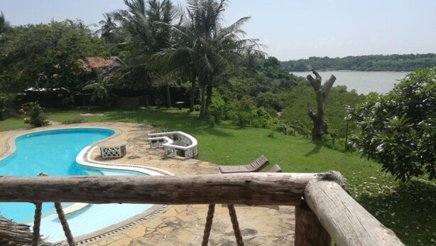 A pool with a stone deck and lounge chairs, surrounded by greenery and a view of a lake. - Cooke Villa (Mtwapa)