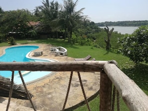 A pool with a stone deck and lounge chairs, surrounded by greenery and a view of a lake. - Cooke Villa (Mtwapa)