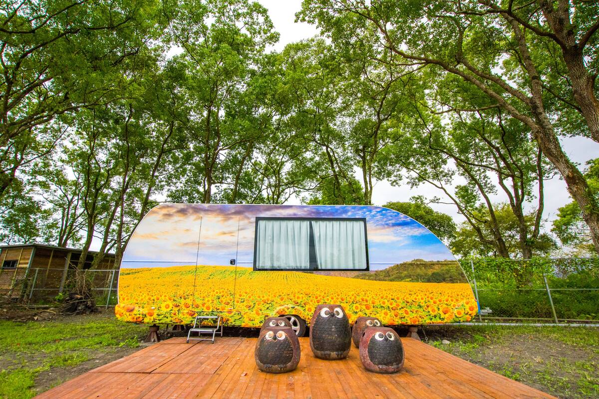 A colorful RV with a sunflower field mural, surrounded by trees.